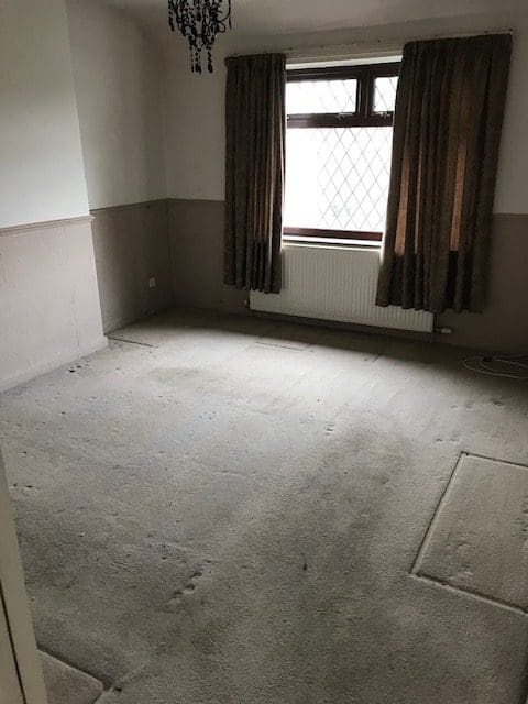 Empty room with worn carpet, dark curtains, and a radiator under the window, showing signs of previous furniture and in need of clearance and cleaning.