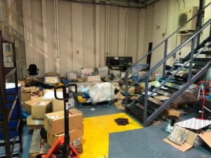 4F7B88F3 0C1E 4D84 9Db6 33E1B08D162A Industrial Clearance A cluttered industrial unit floor covered in stacked boxes, shrink-wrapped pallets, and loose packaging waste, with a pallet jack in the foreground, showing the need for a clearance service.
