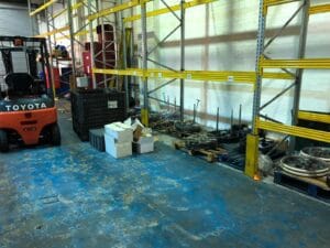 792C6809 41Fc 4B9D 94F6 18C9Cd24032D Industrial Clearance A bright orange toyota forklift parked next to empty pallet racking, with discarded engine parts and debris on wooden pallets and a worn, blue-painted concrete floor.