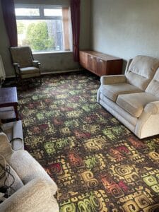 House Clearance Birmingham1 Garden Clearance A cluttered living room with a patterned carpet, two light-colored sofas, an armchair, and a wooden credenza, showing furniture to be cleared from a property.