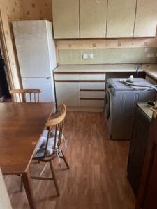 House Clearance Birmingham2 Garden Clearance A kitchen and dining area showing a wooden table and chairs, a washing machine, a large fridge-freezer, and older cabinets, all requiring white goods and furniture removal.