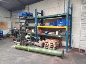 B74974Ba Adbb 4Bee 94Ad 3D22D61261F3 Industrial Clearance A disorganized storage area with blue and yellow industrial racking shelves holding various boxes and plastic totes, with wooden pallets and a large, green metal pipe lying on the floor.