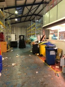 B98F1C78 6Ec2 480E B1Dd Cb341C1E49C3 Industrial Clearance A forklift near a pile of assorted industrial waste, general rubbish, and discarded materials next to a blue "general waste" bin in a large, cluttered warehouse setting.