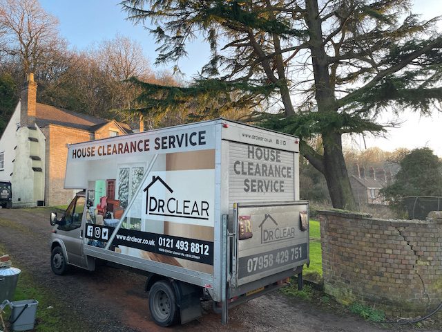 Drclear House Clearance Truck West Midlands Home A long, light grey box truck with 'drclear house clearance service' branding parked on a dirt driveway. The truck has a large graphic of a house outline and contact details including a website and phone numbers. In the background, there's a white house with a brown roof and a tall, mature tree, suggesting a rural or suburban setting under natural light.