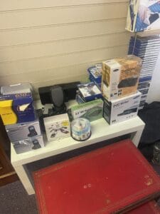 Retail Shop Clearance West Midlands2 Retail Shop Clearance A small white retail display table cluttered with assorted old electronic stock and packaging. Items include boxes for a dvd writer, an omniview kvm switch, a digital mobile phone, a spindle of blank cds, and a small stack of ps4 video game cases. A red, low leather-topped box is in the foreground.