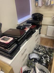 Retail Shop Clearance West Midlands4 Retail Shop Clearance A shop window display area featuring a large stack of discarded laptops and tablets piled on top of a white storage cabinet. A plastic bin on the floor is overflowing with broken mobile phone screens and components, clearly showing electronic repair waste.