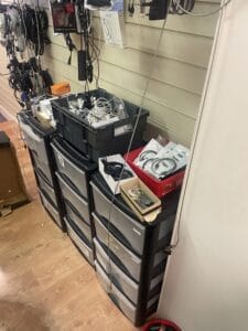 Retail Shop Clearance West Midlands6 Retail Shop Clearance A photo focused on a stack of grey and black plastic drawer units used for storage. The tops of the drawers are littered with small bins and open boxes containing a tangle of various wires, cables, and small electronic components, showing the detail of the parts clearance.