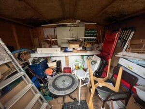 Garage Clearance West Midlands1 Garage Clearance A wide interior view of a cluttered wooden shed or outbuilding. The space is packed with miscellaneous items, including an aluminum ladder, garden furniture (a mosaic-top table, plastic chairs), a red upright step ladder, cardboard boxes, and various tools piled on a workbench across the back wall. The shed roof and walls are made of plywood sheeting.