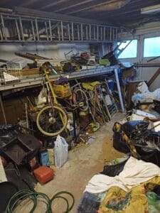 Garage Clearance West Midlands2 Garage Clearance An indoor photo of a very cluttered garage area. A large section of the room is filled with trash bagged in black sacks and loose junk. Resting on top of a pile of debris is a tall extension ladder and several old bicycle frames, including a prominent yellow and red bicycle in the center. Windows are visible high up on the back wall.