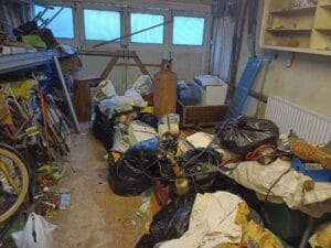 Garage Clearance West Midlands3 Garage Clearance An indoor photo of a messy garage or workshop area. The space is filled with large piles of trash and junk bagged in black plastic sacks, cardboard boxes, and loose items piled up on a table to the right. A large orange gas cylinder stands in the center. The background features light-blue, paned windows across the back wall.