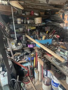 Garage Clearance West Midlands4 Scaled E1762345942553 Garage Clearance A very dense, vertical, close-up shot of a heavily cluttered workshop or garage shelf. The area is overflowing with hand tools, paint cans, various small components, wooden beams, a blue plastic bin overflowing with objects, and electrical cords.