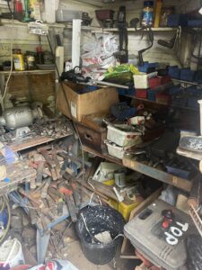 Garage Clearance West Midlands5 Scaled E1762345918799 Garage Clearance A close-up, high-angle, vertical shot of an extremely cluttered space, possibly a corner of a garage. The floor and shelves are completely covered with tools, metal scraps, rusty objects (like tent pegs or spikes), empty cans, plastic crates, wooden planks, and a portable propane tank near the center bottom.