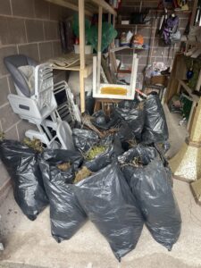 Garage Clearance West Midlands6 Scaled E1762345907880 Garage Clearance A vertical photo showing several large black plastic sacks, tightly packed and lying on a concrete or tiled floor. These bags appear to contain garden waste and debris. Various light-colored plastic items, including baby chairs and furniture parts, are stacked or leaning against a wall next to the bags.
