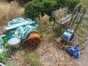 Garden Clearance West Midlands1 Garden Clearance An outdoor shot of garden clutter on a patio or paved area surrounded by dry grass and overgrown weeds. Items include an orange coiled garden hose on a reel, a blue pressure washer unit, a large paint can, a pile of green-and-white striped plastic sheeting, and a dilapidated wooden and wrought-iron garden bench covered in debris in the background.