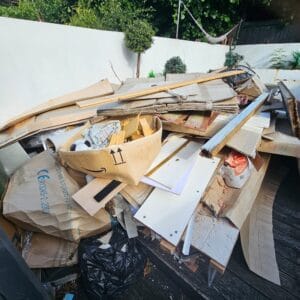 Garden Clearance West Midlands2 Garden Clearance A large, high pile of cardboard boxes, scrap wood, and general packaging waste dominates the frame. A distinctive amazon logo is visible on a partially filled box in the center. The waste is piled on what appears to be dark decking or a patio, set against a bright white garden wall with small, trimmed shrubs in the background.