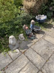 Garden Clearance West Midlands4 Garden Clearance A sunlit, close-up view of a garden path made of large, square paving stones. A line of various old garden ornaments rests on the stones near a border of green foliage. The ornaments include a concrete owl, a small rabbit, and two garden gnomes with a wheelbarrow and a planter.