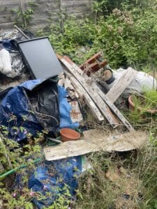 Garden Clearance West Midlands5 Garden Clearance A tight, vertical close-up of a dense pile of garden waste and scrap materials engulfed by tall weeds and greenery. The pile consists of broken wooden planks, a rusted metal piece, black and blue plastic sheeting (tarpaulin), a black plastic tray, and several small terracotta plant pots.