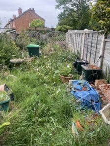 Garden Clearance West Midlands6 Garden Clearance A wide shot of a heavily overgrown, messy back garden. Tall weeds, dandelions, and long grass cover the ground. The area contains two green council bins overflowing with waste, a birdbath, several empty terracotta pots, a pile of rubble and broken bricks, and a discarded blue tarpaulin. The garden is enclosed by a wooden fence, with the back of a brick terraced house visible beyond.