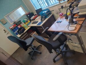 Office Clearance West Midlands1 Office Clearance A slanted view of a typical messy office corner with two adjacent desks. The desks are covered with paperwork, binders, landline phones, and desktop computers. Two black wheeled office chairs are pushed against the desks. The walls have blue textured wallpaper, and the