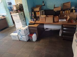Office Clearance West Midlands2 Office Clearance A view of a desk area ready for clearance. The desk surface is covered with several open cardboard boxes and a phone, and it is flanked by dark brown metal filing cabinets. On the floor to the left, stacks of white and blue lyreco archive boxes are piled up next to a bottle of liquid, indicating the office contents are being packed up or prepared for removal.