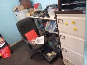 Office Clearance West Midlands3 Office Clearance A photo focusing on a desk chair and a white, four-drawer filing cabinet. The chair has a red folder resting on the seat, and a black box is on the floor nearby. The office has blue textured wallpaper. The filing cabinet has yellow magnetic notes and small labels stuck on the drawers.