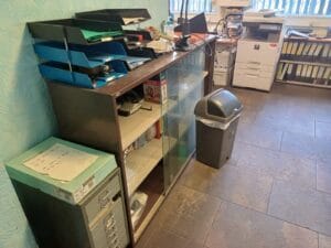 Office Clearance West Midlands4 Office Clearance A picture of a cluttered area featuring a glass-fronted metal storage cabinet. The cabinet holds files and supplies, and on top sit several blue and black plastic paper trays filled with documents. To the left is a grey stack of flat file drawers, and in the background is a large, white office photocopier/printer machine and shelving full of black ring binders. A grey plastic trash can is in the centre.