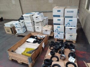 Office Clearance West Midlands5 Office Clearance An industrial setting, likely a warehouse floor, with large quantities of inventory and archived documents. Several stacks of lyreco and impega archive boxes are piled on wooden pallets. In the foreground, a wooden crate holds various black rubber and metal industrial components, seals, and rings, indicating a clearance involving specialized business stock.