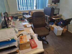 Office Clearance West Midlands6 Office Clearance A wide-angle shot of a very messy office desk dominated by a dark grey, high-back ergonomic chair. The desk is completely covered with papers, a newspaper, a partially eaten plate of food, a box of toffifee, and a desktop monitor. In the background, two computers and multiple monitors sit on a small, cluttered stand near a window with vertical blue blinds. The room has dark tiled flooring and blue textured wallpaper.