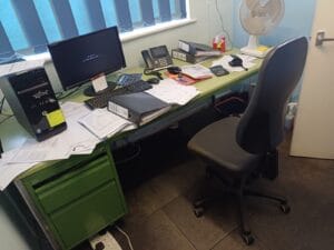 Office Clearance West Midlands7 Office Clearance A slightly neater, but still cluttered, office desk. The desk surface is a pale green, with matching green metal filing drawers on the left. Scattered papers, black folders, a calculator, a landline phone, and a computer tower are on the desk. A white oscillating fan is visible on the right, and a black office chair is pulled up to the desk.