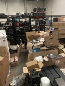 Warehouse Clearance West Midlands1 Warehouse Clearance A cluttered view inside a warehouse aisle. On the left, several black metal shelving units are packed with black electronic components and computer cases, some stacked precariously. The floor and lower shelving are blocked by a large, chaotic pile of open brown cardboard boxes, many marked "fragile," suggesting the clearance involves sorting through stock.